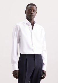 Camicia bianca a maniche lunghe con colletto button-down, realizzata in tessuto morbido, abbinata a pantaloni navy su misura. Dettaglio di texture sottile.