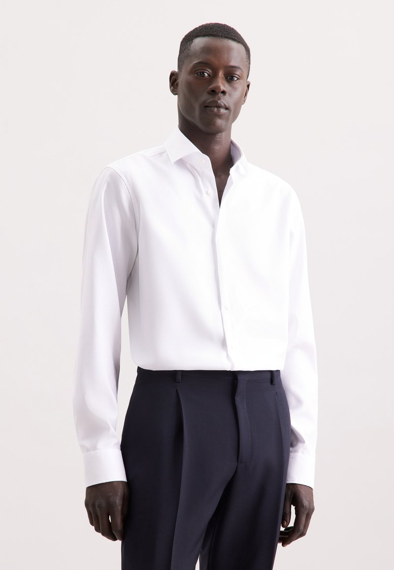 Camicia bianca a maniche lunghe con colletto button-down, realizzata in tessuto morbido, abbinata a pantaloni navy su misura. Dettaglio di texture sottile.