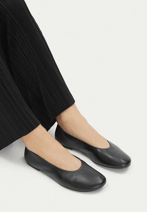 Osoba nosząca czarne, skórzane, płaskie buty z okrągłymi noskami oraz czarne plisowane szerokie spodnie na jasnym tle.