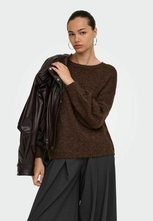 Jeune femme aux cheveux tirés en arrière, portant de grandes boucles d’oreilles créoles, un pull marron duveteux, un pantalon large gris foncé, et tenant une veste en cuir foncée sur l’épaule.