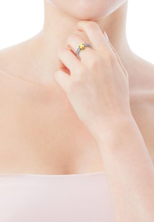 Silber- und Goldring mit einem gelben Akzent in floraler Gestaltung, mit strukturiertem Band und glatter Oberfläche. An einer Hand vor einem hellen Hintergrund getragen.