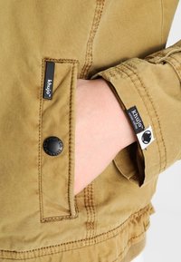 Kaki jacka med knappstängning, sydda fickor och loggade taggar på ärmen. Tyget har en texturerad och slitstark finish.