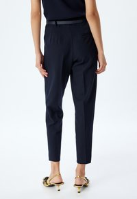 Pantalones de traje en color navy con un corte slim, que presentan una textura suave, trabillas para el cinturón y un bolsillo trasero. Combinados con sandalias doradas de tacón.