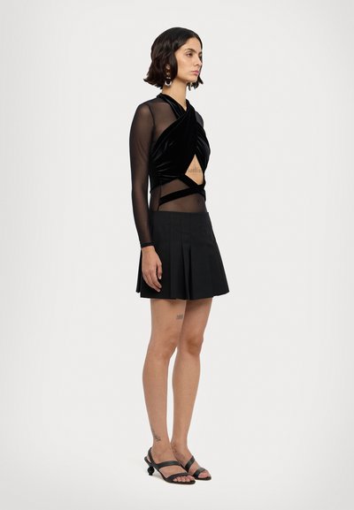 Sort velour langærmet top med uitsnit og mesh-sektioner, parret med en plisseret sort miniskirt og snørede sorte sandaler.