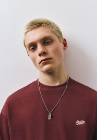 Jeune personne aux cheveux blonds courts portant un t-shirt bordeaux et un collier pendentif en argent, regardant légèrement vers le bas sur un fond uni.