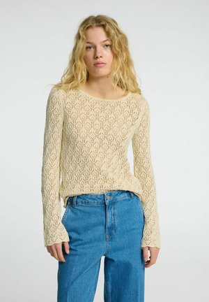 Beige Strickpullover mit durchgehendem Rautenmuster, langen Ärmeln und weit ausgeschnittenem Rundhalsausschnitt. Kombiniert mit hoch geschnittenen blauen Jeans.