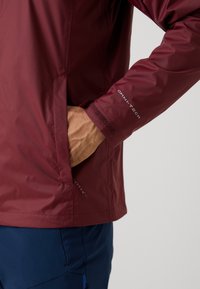 Persoon draagt een wijnrode waterdichte jas met "Omni-Tech" op de mouw, hand in de zijzak gestoken, gecombineerd met marineblauwe broek.