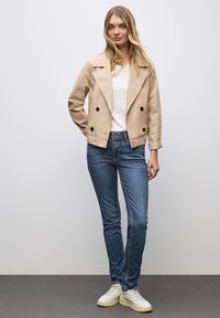 Beżowa krótka kurtka z podwójnymi guzikami, zestawiona z niebieskimi dżinsami. Modelka nosi białą bluzkę i białe trampki.