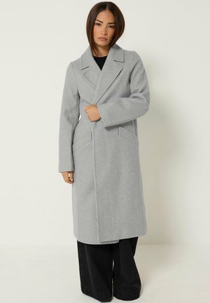 Femme portant un long manteau en laine gris clair sur un pantalon noir à jambes larges, debout devant un fond clair uni.
