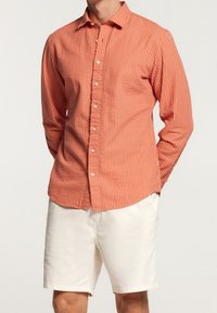 Red and white striped button-up shirt made of textured fabric, paired with cream-colored shorts. Features a collar and long sleeves.