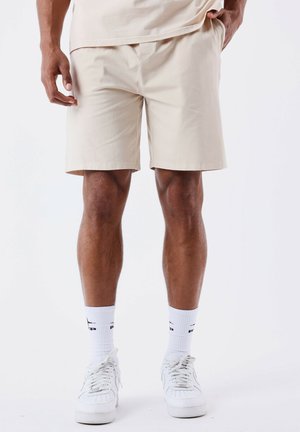 Pantalones cortos de algodón beige con un ajuste relajado, diseño liso y bolsillos laterales, combinados con zapatillas blancas y calcetines blancos con logo.