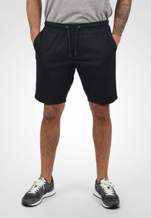 Shorts noirs avec une taille élastique et un cordon de serrage, dotés de poches latérales. Portés avec des chaussures de sport grises et marine.