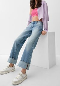 Mujer apoyada en un bloque blanco, vestida con jeans de pierna ancha remangados, zapatillas blancas, un top corto rosa y una camisa morada abotonada sobre un hombro.