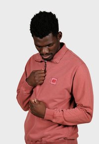 Sweatshirt couleur rouille avec un col zippé, arborant un patch logo rouge. Le tissu semble doux avec des poignets et un ourlet côtelés.