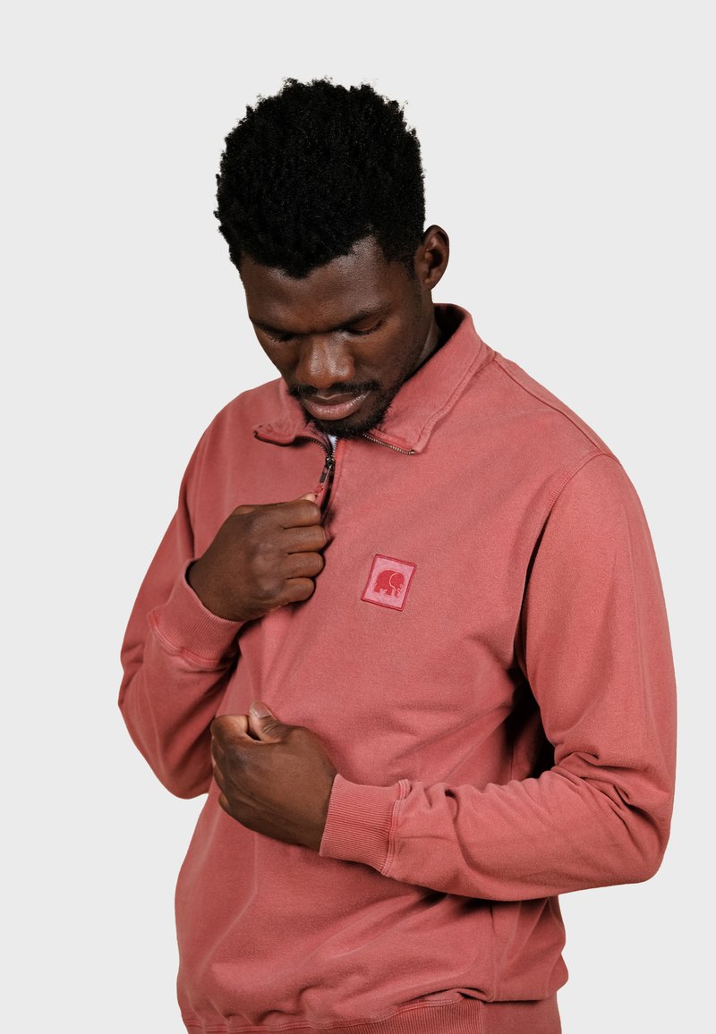 Sweatshirt couleur rouille avec un col zippé, arborant un patch logo rouge. Le tissu semble doux avec des poignets et un ourlet côtelés.