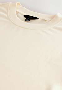 Camiseta de algodón crema con cuello redondo, mangas cortas y una textura suave. Presenta una etiqueta que indica la marca y la talla en el interior del cuello.