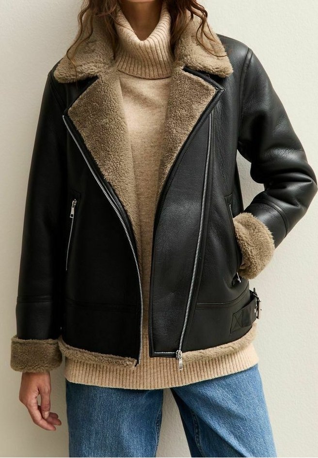 Femme portant une veste d'aviateur en cuir noir avec doublure en peau retournée beige, un pull à col roulé beige et un jean bleu, debout contre un mur clair.