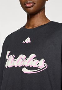 Μαύρο βαμβακερό t-shirt με μεγάλο λευκό και ροζ λογότυπο "adidas" σε παιγνιώδη γραμματοσειρά, με γεωμετρικό διακοσμητικό πάνω από το λογότυπο.