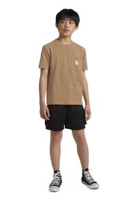 Ruda trumpo rankovės marškinėliai su kišene ir logo, derinami su juodomis tinklinėmis šortais ir juodais aukštakulniais sportbačiais.