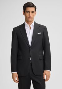 Giacca nera con rever a punta, due bottoni e due tasche. Abbinata a una camicia bianca con colletto a punta.