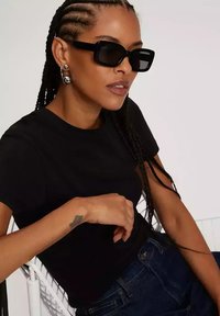 Czarna koszulka, prostokątne czarne okulary przeciwsłoneczne, warkocze i dżinsowe spodnie. Dodatki obejmują okrągłe kolczyki oraz subtelny tatuaż na ramieniu.