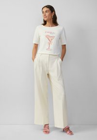 Wit T-shirt met een roze cocktailafbeelding en tekst, gecombineerd met hooggewaadde, crèmewitte wijde broek. Lichtroze sandalen met bandjes.