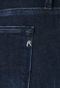 El tejido de mezclilla de color azul oscuro presenta un pequeño acento plateado en forma de "R" en el bolsillo trasero. Un distintivo ribete resalta los bordes del bolsillo.