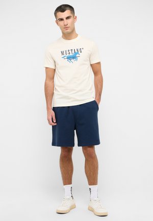 Beige cotton t-shirt with blue "MUSTANG" logo and graphic, paired with navy blue shorts. Model wearing white sneakers and ankle socks.