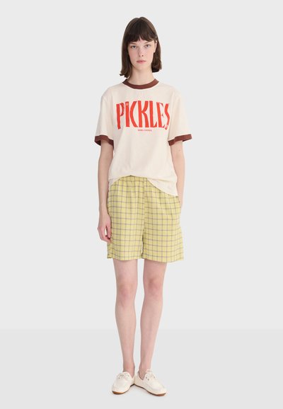 Femme debout portant un t-shirt crème avec le mot "PICKLES" en rouge, un short à carreaux jaunes, et des baskets blanches sur un fond uni.