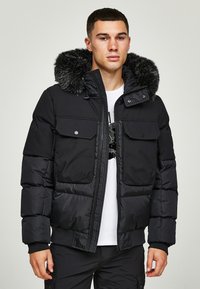 Svart pufferjakke med avtagbar faux fur hette, to brystlommer, glidelås, ribbestrikkede mansjetter og en jevn tekstur.