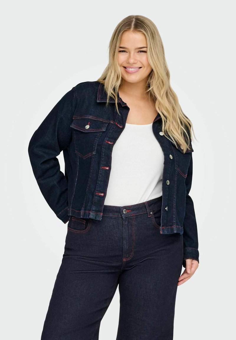 Leende kvinna iklädd en mörk denimjacka över en vit topp och matchande mörka denimjeans med röda sömmar, framför en enfärgad bakgrund.