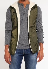 Grön vadderad väst med sherpa-foder, bärs över en grå långärmad henley. Har dragkedjestängning och sidofickor.