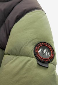 Manga de chaqueta acolchada en verde olivo y gris oscuro, con un parche redondo de logo en negro y rojo que presenta gráficos de montañas y detalles de texto.