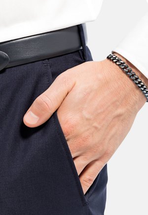 Pantalones de navy de tejido suave; un cinturón negro de acabado elegante; una mano reposando en el bolsillo lateral; pulsera de cadena de plata en la muñeca.