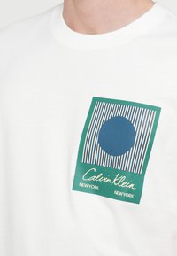 White cotton t-shirt featuring a green rectangular logo with a blue circle, thin vertical stripes, and 'Calvin Klein’ text in a cursive font.