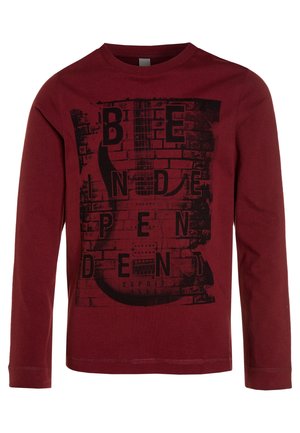 Chemise manches longues marron avec un graphisme noir de guitare électrique et un texte fragmenté épelant "Independent" sur un fond de mur en briques.