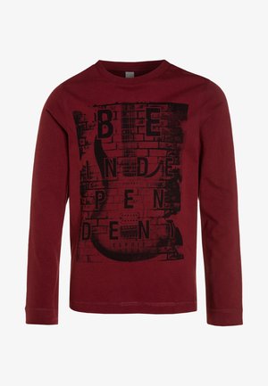 Chemise manches longues marron avec un graphisme noir de guitare électrique et un texte fragmenté épelant "Independent" sur un fond de mur en briques.