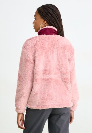 Rosa fluffy fleece-jakke med høy krage, myk tekstur og lange ermer, båret over mørke bukser. Har en avslappet passform og ingen synlige lukkinger.