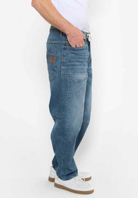 Jeans de mezclilla azul claro con un ajuste relajado, costuras visibles, cinco bolsillos y una etiqueta de cuero en la parte trasera. Llevados con zapatillas blancas.