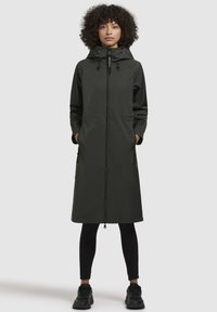 Cappotto lungo impermeabile verde scuro con zip, cappuccio regolabile, due tasche laterali e taglio dritto, abbinato a leggings neri e scarpe.