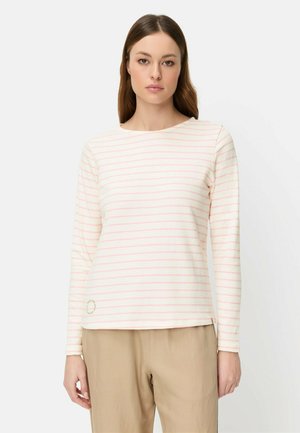 Vrouw die een wit shirt met lange mouwen draagt met dunne roze horizontale strepen en beige broek, staand tegen een effen lichte achtergrond.