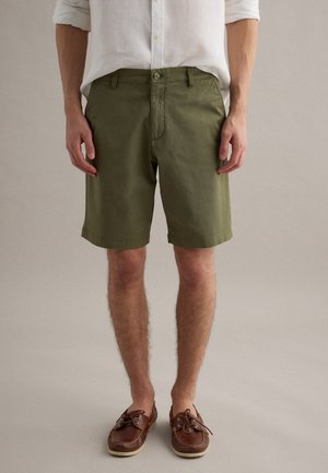 Man draagt olijfgroene knielange shorts, een wit opgerolde lange mouwen overhemd en bruine leren bootschoenen, staand tegen een effen achtergrond.