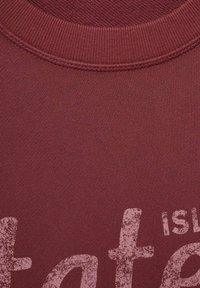 Zvětšený pohled na tkaninu v bordó barvě s žebrovým vzorem a částečně vybledlým světle růžovým textem, který ukazuje písmena "is" a "te".