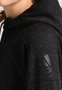 Sweat à capuche noir à texture tachetée, avec un logo Adidas noir sur la manche et une capuche à cordon. Tissu doux, coupe décontractée.