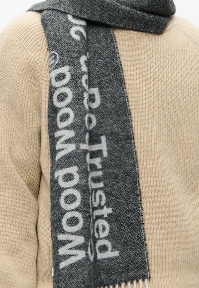 Hall kivivõrkline sall, millel on julged valged tekstid, narmastega servad ja pehme tekstuur, riputatud beežile kootud sviitrile.