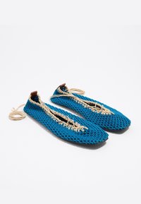 Blauwe geweven stoffen schoenen met een platte vorm, voorzien van een gevlochten touw in natuurlijke kleuren en een bruine leren accent bij de hiel.