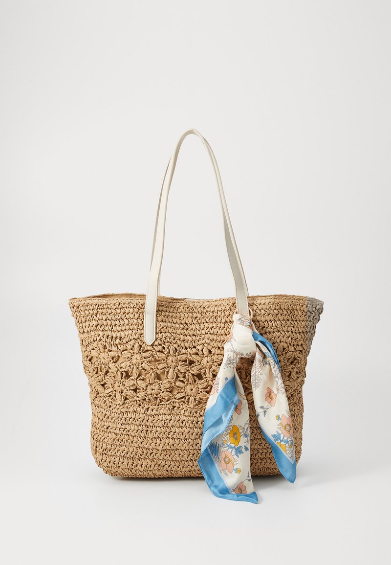 Saco de palha entrelaçado na cor natural com alças brancas e um lenço de seda com padrão floral decorativo amarrado de um lado.