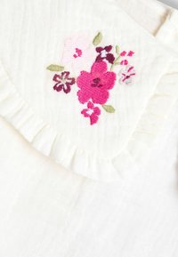 Țesătură albă cu un guler ondulat, decorată cu broderie florală în nuanțe de roz, mov și verde. Textura pare moale și texturată.