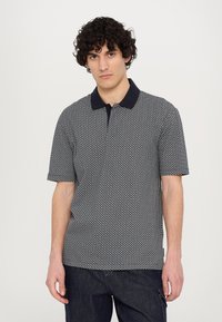 Kortermet polo skjorte i svart og hvitt geometrisk mønster, med en mørkeblå krage og strukturert stoff. Har en rettkutt design.
