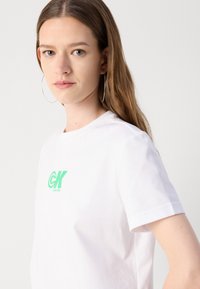 Balta medvilnės marškinėliai su žaliu Calvin Klein logotipu krūtinėje. Trumpomis rankovėmis ir apvalia iškirpte. Lygus audinys.